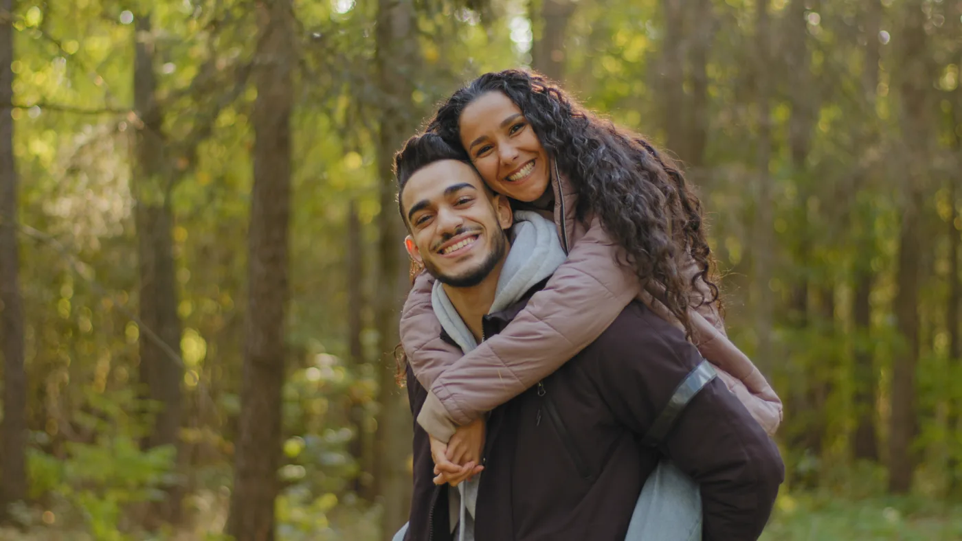 Two people smile during a piggyback ride in a forest.